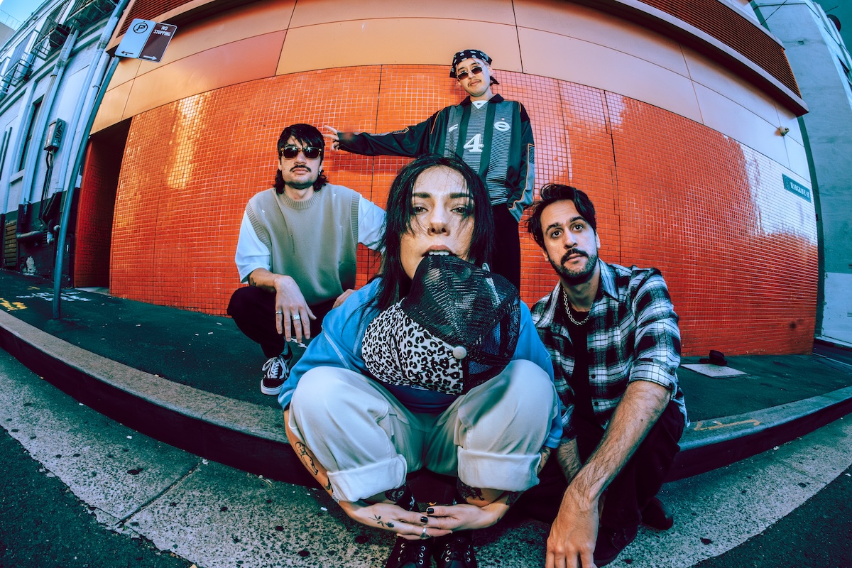 Australian pop-punk band Stand Atlantic posing outdoors against an orange wall. Bonnie Fraser crouches in front, biting a leopard-print cap.
